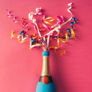 Champagne bottle with colorful party streamers on pink background. Flat lay
